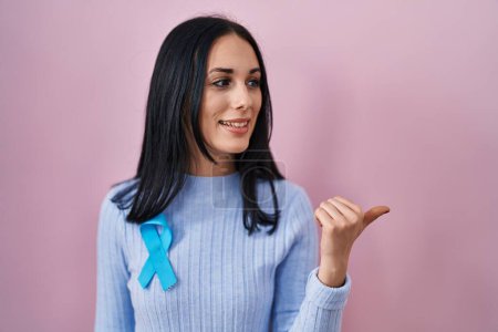 Foto de Mujer hispana con cinta azul apuntando con el pulgar hacia el costado sonriendo feliz con la boca abierta - Imagen libre de derechos