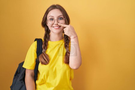 Téléchargez les photos : Jeune femme caucasienne portant un sac à dos étudiant sur fond jaune pointant du doigt vers le visage et le nez, souriante et joyeuse. concept de beauté - en image libre de droit