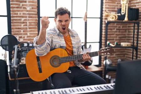 Photo for Young hispanic man playing classic guitar at music studio annoyed and frustrated shouting with anger, yelling crazy with anger and hand raised - Royalty Free Image