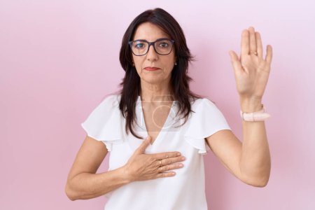 Téléchargez les photos : Femme hispanique du Moyen Âge portant un t-shirt blanc décontracté et des lunettes jurant avec la main sur la poitrine et la paume ouverte, faisant un serment de fidélité - en image libre de droit