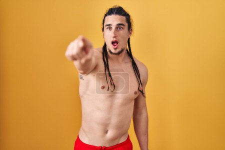 Téléchargez les photos : Homme hispanique avec les cheveux longs debout torse nu sur fond jaune pointant avec le doigt surpris devant, bouche ouverte expression étonnée, quelque chose sur le devant - en image libre de droit
