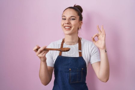 Photo for Young hispanic girl wearing professional cook apron holding wood spoon smiling and laughing hard out loud because funny crazy joke. - Royalty Free Image