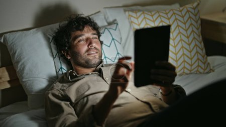 Téléchargez les photos : Young hispanic man using touchpad lying on bed at bedroom - en image libre de droit