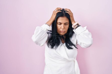 Foto de Mujer hispana madura de pie sobre fondo rosa que sufre de dolor de cabeza desesperada y estresada porque el dolor y la migraña. manos en la cabeza. - Imagen libre de derechos