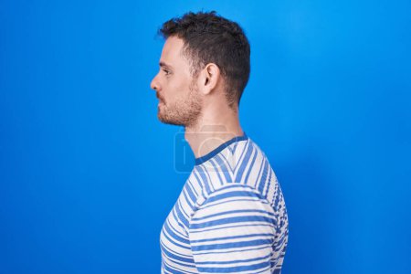 Photo for Young hispanic man standing over blue background looking to side, relax profile pose with natural face and confident smile. - Royalty Free Image