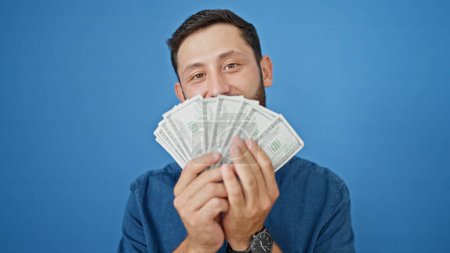 Téléchargez les photos : Jeune homme hispanique souriant bouche couverture confiante avec des dollars sur fond bleu isolé - en image libre de droit