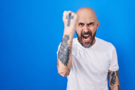 Téléchargez les photos : Homme hispanique avec des tatouages debout sur fond bleu en colère et fou levant le poing frustré et furieux tout en criant de colère. rage et concept agressif. - en image libre de droit
