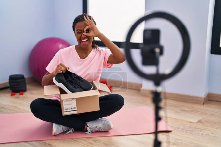 Foto de Joven afroamericano con trenzas grabando vlog tutorial con smartphone en el gimnasio sonriendo feliz haciendo ok signo con la mano en el ojo mirando a través de los dedos - Imagen libre de derechos