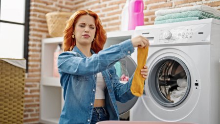 Téléchargez les photos : Jeune rousse femme laver les vêtements avec une expression détendue à la buanderie - en image libre de droit