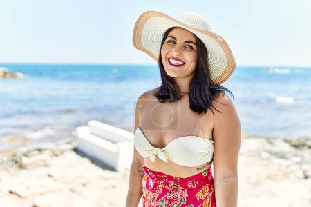Foto de Joven mujer hispana hermosa turista sonriendo confiado usando bikini y sombrero de verano en la playa - Imagen libre de derechos