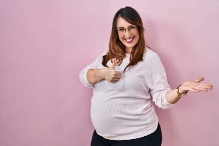 Téléchargez les photos : Femme enceinte debout sur fond rose montrant la main de la paume et faisant un geste correct avec les pouces levés, souriant heureux et joyeux - en image libre de droit