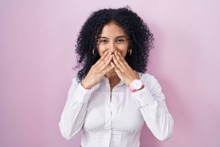 Foto de Mujer hispana con el pelo rizado de pie sobre fondo rosa riendo y riéndose avergonzada cubriendo la boca con las manos, chismes y concepto de escándalo - Imagen libre de derechos