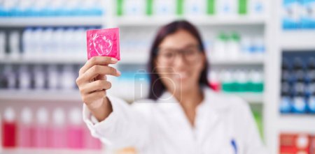 Foto de Joven bella mujer hispana farmacéutica sonriendo confiada sosteniendo el condón en la farmacia - Imagen libre de derechos