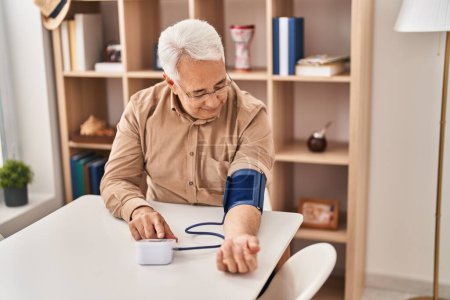 Foto de Hombre mayor usando tensiómetro sentado en la mesa en casa - Imagen libre de derechos
