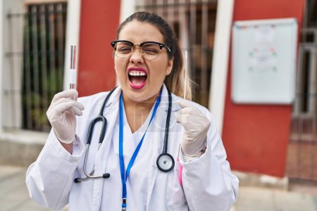 Foto de Joven doctora hispana haciendo prueba nasal de infección por coronavirus enojada y loca gritando frustrada y furiosa, gritando de rabia. rabia y concepto agresivo. - Imagen libre de derechos