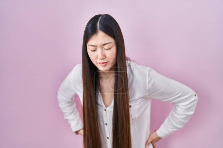 Téléchargez les photos : Jeune femme chinoise debout sur fond rose souffrant de maux de dos, toucher le dos avec la main, douleur musculaire - en image libre de droit