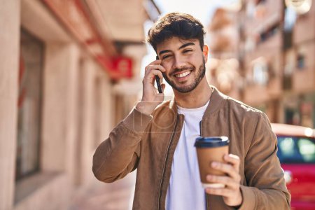Téléchargez les photos : Jeune homme hispanique parlant sur le smartphone boire du café dans la rue - en image libre de droit