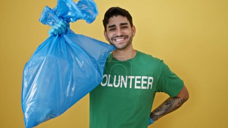 Foto de Joven hispano voluntario usando guantes sosteniendo bolsa de basura sobre aislado fondo amarillo - Imagen libre de derechos