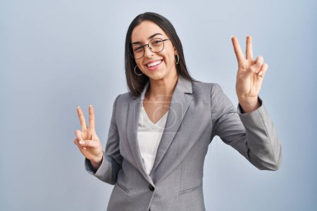 Foto de Mujer de negocios hispana con gafas sonrientes mirando a la cámara mostrando los dedos haciendo la señal de victoria. número dos. - Imagen libre de derechos
