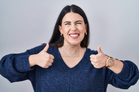 Foto de Young brunette woman standing over isolated background success sign doing positive gesture with hand, thumbs up smiling and happy. cheerful expression and winner gesture. - Imagen libre de derechos