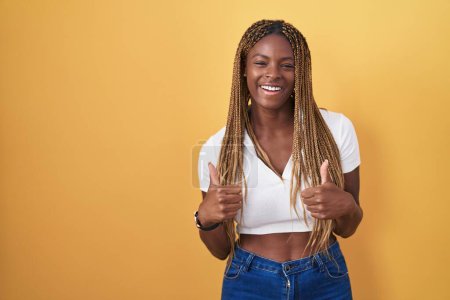 Téléchargez les photos : Femme afro-américaine avec les cheveux tressés debout sur fond jaune signe de succès faisant geste positif avec la main, pouces levés souriant et heureux. expression joyeuse et geste gagnant. - en image libre de droit
