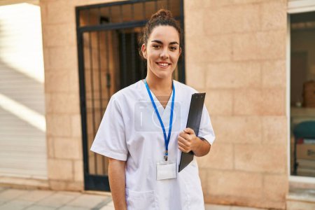 Téléchargez les photos : Jeune belle hispanique femme physiothérapeute souriant confiant tenant liste de contrôle à la rue - en image libre de droit