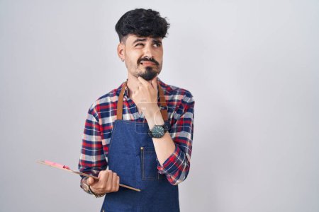 Foto de Joven hispano con barba vistiendo delantal de camarero sosteniendo portapapeles pensando preocupado por una pregunta, preocupado y nervioso con la mano en la barbilla - Imagen libre de derechos