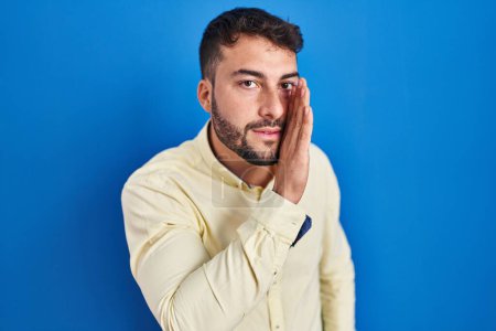 Photo for Handsome hispanic man standing over blue background hand on mouth telling secret rumor, whispering malicious talk conversation - Royalty Free Image