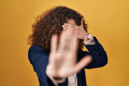Téléchargez les photos : Femme hispanique avec les cheveux bouclés debout sur fond jaune couvrant les yeux avec les mains et faisant arrêter geste avec expression triste et peur. concept embarrassé et négatif. - en image libre de droit