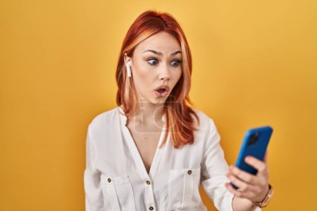 Téléchargez les photos : Jeune femme caucasienne utilisant un smartphone portant des écouteurs effrayés et étonnés avec la bouche ouverte pour la surprise, visage incrédule - en image libre de droit