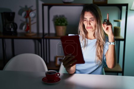 Photo for Young hispanic woman using touchpad sitting on the table at night pointing up looking sad and upset, indicating direction with fingers, unhappy and depressed. - Royalty Free Image
