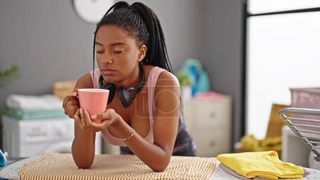 Téléchargez les photos : Femme afro-américaine buvant du café appuyé sur la planche à repasser fatigué à la buanderie - en image libre de droit