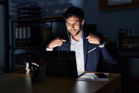 Foto de Hombre latino guapo trabajando en la oficina por la noche mirando confiado con sonrisa en la cara, señalándose con los dedos orgullosos y felices. - Imagen libre de derechos