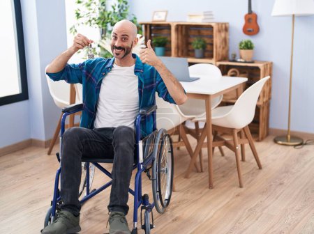 Téléchargez les photos : Homme hispanique avec barbe assis sur fauteuil roulant tenant de nouvelles clés de la maison souriant heureux et positif, pouce levé faire excellent et signe d'approbation - en image libre de droit