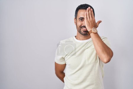 Téléchargez les photos : Hispanic man with beard standing over isolated background covering one eye with hand, confident smile on face and surprise emotion. - en image libre de droit