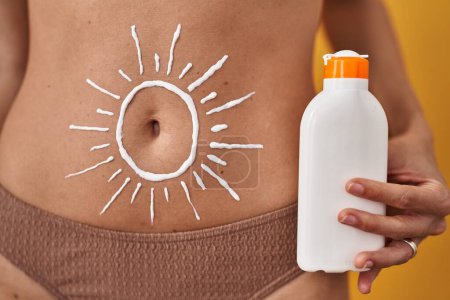 Téléchargez les photos : Jeune femme en bikini avec lotion solaire dessiner sur le ventre sur fond jaune isolé - en image libre de droit