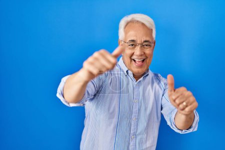 Foto de Hombre mayor hispano con gafas aprobando hacer gesto positivo con la mano, pulgares arriba sonriendo y feliz por el éxito. gesto ganador. - Imagen libre de derechos
