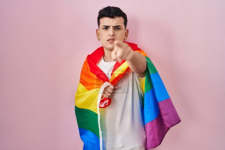 Photo for Non binary person holding rainbow lgbtq flag pointing with finger to the camera and to you, confident gesture looking serious - Royalty Free Image