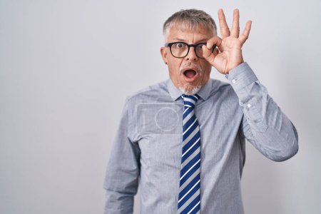 Foto de Hombre de negocios hispano con el pelo gris usando gafas haciendo un gesto bien sorprendido con la cara sorprendida, los ojos mirando a través de los dedos. expresión incrédula. - Imagen libre de derechos