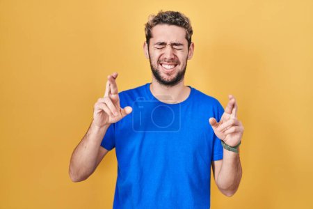 Téléchargez les photos : Homme hispanique avec barbe debout sur fond jaune geste doigt croisé souriant avec espoir et les yeux fermés. chance et concept superstitieux. - en image libre de droit
