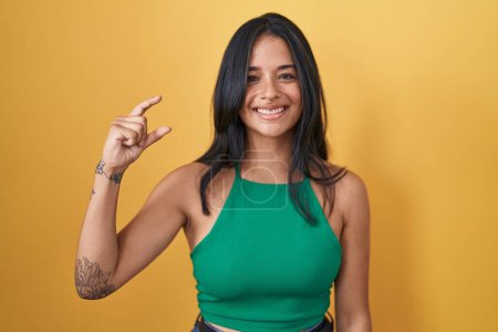 Photo for Brunette woman standing over yellow background smiling and confident gesturing with hand doing small size sign with fingers looking and the camera. measure concept. - Royalty Free Image