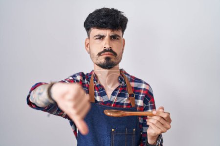 Téléchargez les photos : Jeune homme hispanique avec barbe portant tablier dégustation de nourriture tenant cuillère en bois avec le visage en colère, signe négatif montrant aversion avec les pouces vers le bas, concept de rejet - en image libre de droit