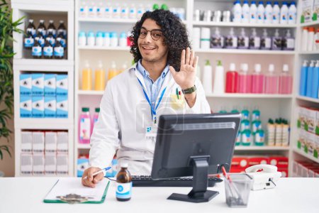 Téléchargez les photos : Homme hispanique aux cheveux bouclés travaillant à la pharmacie pharmacie renonçant à dire bonjour heureux et souriant, geste de bienvenue amical - en image libre de droit