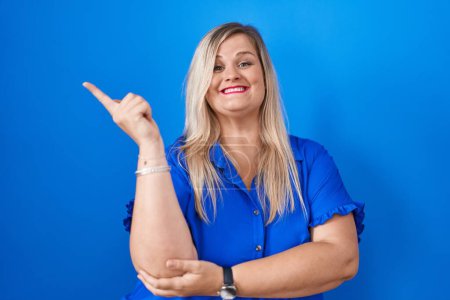 Photo for Caucasian plus size woman standing over blue background with a big smile on face, pointing with hand and finger to the side looking at the camera. - Royalty Free Image