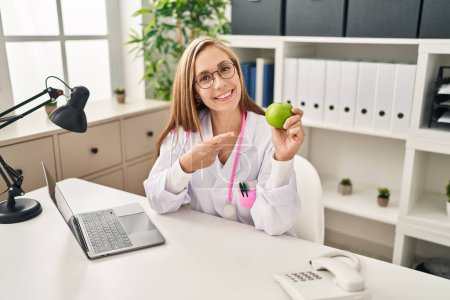 Foto de Joven doctora rubia sosteniendo manzana verde en la clínica sonriendo feliz señalando con la mano y el dedo - Imagen libre de derechos