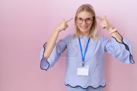Téléchargez les photos : Jeune femme d'affaires caucasienne portant une carte d'identité souriant pointant vers la tête avec les deux doigts des mains, grande idée ou pensée, bonne mémoire - en image libre de droit
