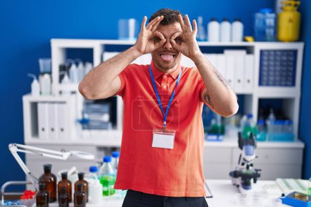 Photo for Young hispanic man working at scientist laboratory doing ok gesture like binoculars sticking tongue out, eyes looking through fingers. crazy expression. - Royalty Free Image