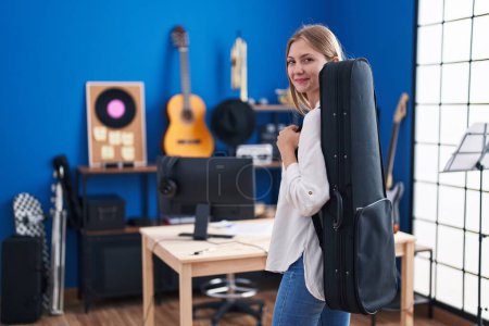 Photo for Young caucasian woman artist smiling confident holding guitar case at music studio - Royalty Free Image