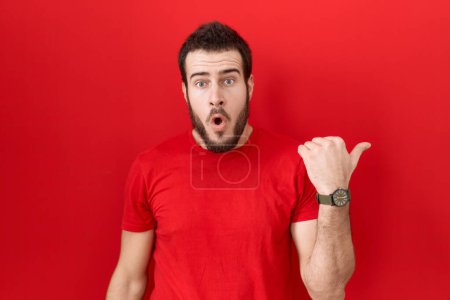 Téléchargez les photos : Jeune homme hispanique portant un t-shirt rouge décontracté surpris pointant du doigt sur le côté, bouche ouverte expression étonnante. - en image libre de droit