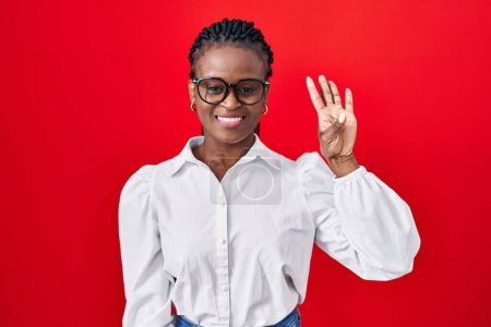 Foto de Mujer africana con trenzas de pie sobre fondo rojo mostrando y señalando hacia arriba con los dedos número cuatro mientras sonríe confiado y feliz. - Imagen libre de derechos
